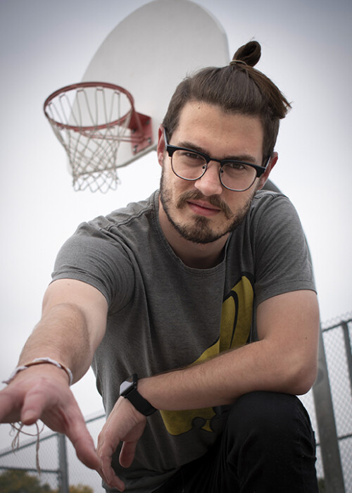 man wearing glasses positioned on one knee extending his right hand towards the camera while his left hand rests on his bent left knee. as background the image shows a basketball hoop and parts of the fence surrounding the court.