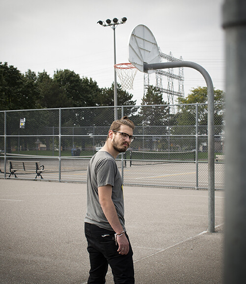 man wearing glasses entering a basketball court looking back over his right shoulder directly at the camera. The image shows a view of part of the court with the basketball basket. Part of the iron gate which serves as entrance to the court is shown in the foreground framing the man who appears in the center