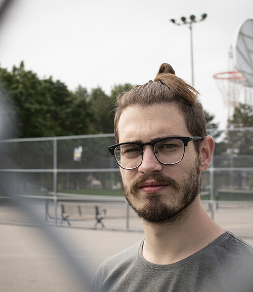 head and shoulders image of a man wearing glasses looking directly into the camera through a fence with a basketball court in a blurred background. Features of the fence and basketball hoop are displayed.